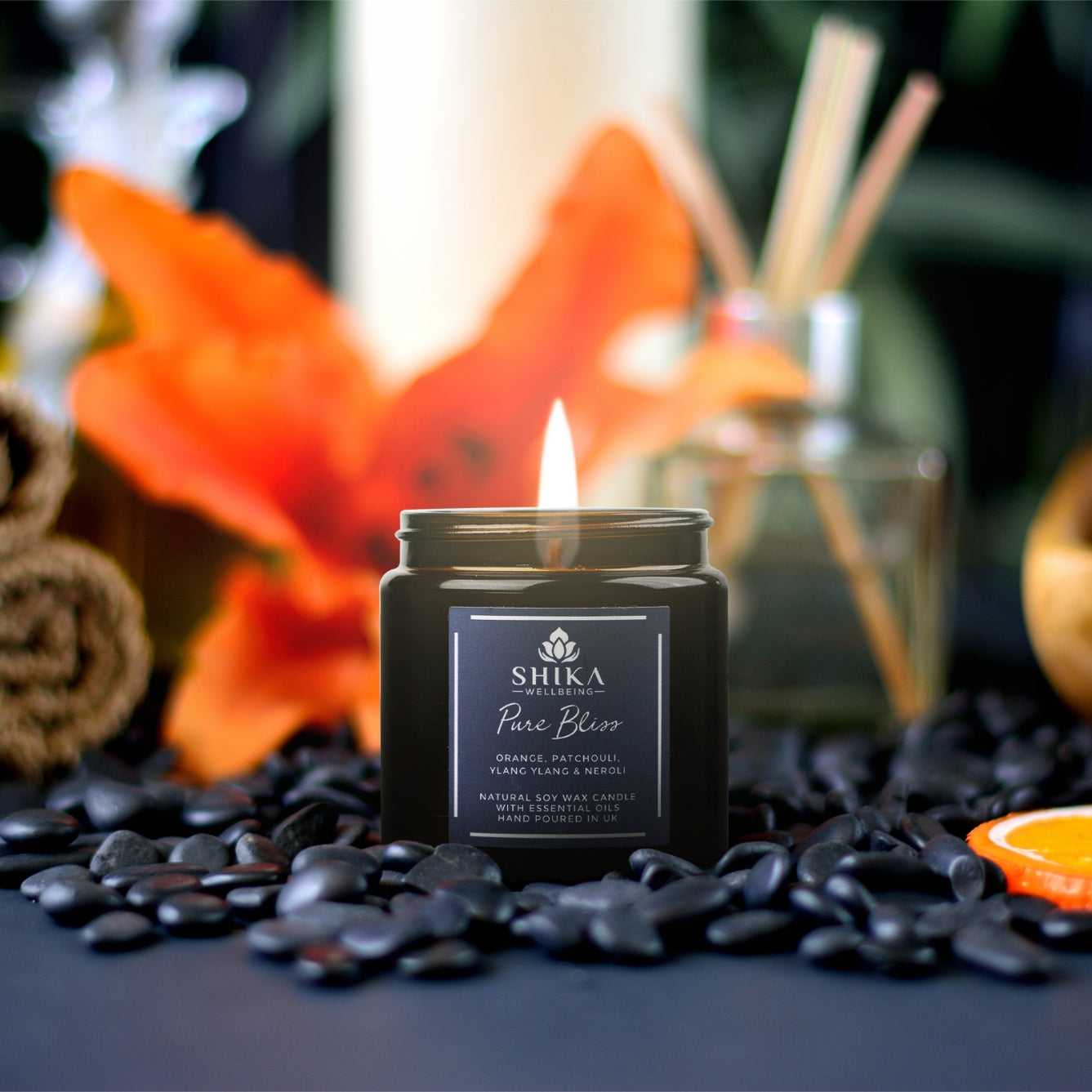 Image of a Shika Wellbeing Pure Bliss candle surrounded by stones. In the background there is an orange flower, a reed diffuser and a slice of orange